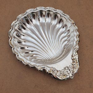 Könnte beinhalten: Eine silberfarbene Metallschale in Form einer Jakobsmuschel. Das Innere weist tiefe, strahlenförmige Rillen auf, während der Griffbereich mit kunstvollen, erhabenen Schnörkeln und blumenartigen Prägungen entlang der Ränder verziert ist.