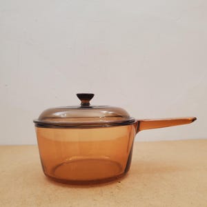 Puede incluir: Un cazo de cristal de color ámbar translúcido con su tapa de cristal a juego. El utensilio de cocina cuenta con un mango largo que se extiende desde un lado y un pequeño pomo de color marrón oscuro centrado en la tapa. El cristal es liso.