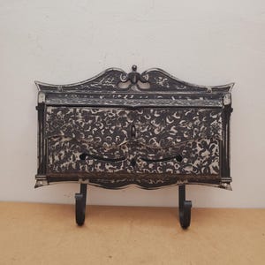 May include: An antique-style mailbox with a black and white floral pattern. The mailbox has two compartments and two hooks below. The top of the mailbox has an ornate, curved design.