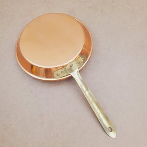 Può includere: Una padella rotonda in rame con finitura metallica lucida e un lungo manico piatto in ottone fissato lateralmente con tre rivetti visibili. La padella è mostrata dal lato inferiore, mostrando una superficie liscia e riflettente.