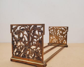 Puede incluir: Un atril extensible de latón con dos paneles finales rectangulares que presentan intrincados patrones florales calados. Las dos piezas de los extremos están conectadas por tres varillas horizontales de latón para ajustar la longitud.