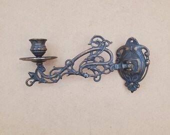 Puede incluir: Un candelabro de pared antiguo con un acabado de bronce oscuro ornamentado. El candelabro presenta una pequeña copa para la vela, un brazo decorativo con detalles florales y de volutas, y una placa trasera para el montaje.