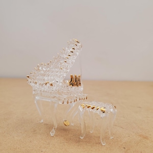 Puede incluir: Un piano de cola en miniatura de cristal transparente con un taburete a juego. El piano y el taburete están adornados con detalles dorados. Las teclas del piano son visibles, y el diseño es delicado y detallado.