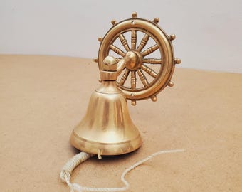 May include: A brass ship's bell with a helm design. The bell is gold-colored and has a rope pull. The helm is attached to the top of the bell. The bell is sitting on a brown surface.