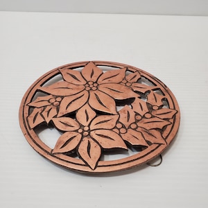 May include: A copper-coloured metal trivet with a floral design featuring poinsettia flowers and holly leaves.