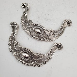 May include: A pair of silver-coloured drawer pulls with elaborate detailing. Each pull has a curved shape with floral and scroll patterns. An oval, raised central section is visible on each. These are likely vintage or antique.