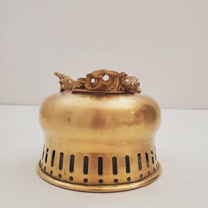 May include: A brass oil lamp burner with a decorative top and vertical slots around the base. The top has an ornate design. The lamp has a polished, golden finish, typical of antique lighting.