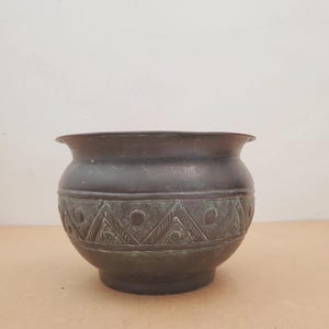 Puede incluir: Un cuenco de metal en tono bronce oscuro con cuerpo redondeado y borde acampanado. El exterior presenta una banda horizontal decorada con un patrón repetitivo de triángulos grabados, texturas punteadas y hendiduras circulares.