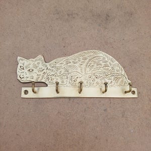 May include: A brass wall-mounted hook rack shaped like a cat with three kittens. The metal piece features an engraved fur texture and five individual hooks attached to a horizontal base bar with two screw holes for mounting on a wall surface.