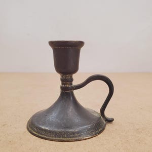 Puede incluir: Un pequeño candelero de estilo antiguo hecho de metal oscuro. Presenta una base ancha y circular, un cuerpo acampanado y una pequeña copa para sostener una vela. Un asa curva se extiende desde el lateral.