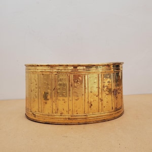 May include: A cylindrical brass container featuring a vertical ribbed pattern along its exterior surface. The metal shows signs of age with visible patina, small dark spots, and surface wear throughout the gold-toned finish of the vessel.
