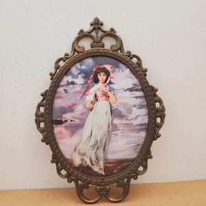 Può includere: Una decorazione da parete ovale con il ritratto di una persona che indossa un abito bianco e una cuffia rosa, su uno sfondo di cielo nuvoloso. L'opera è racchiusa in una cornice metallica color bronzo con volute e dettagli decorativi.