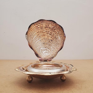 May include: A silver-toned metal trinket box shaped like an open seashell. The interior features a textured, rippled pattern, while the base is supported by three small, rounded ball feet. A circular, smooth silver-toned insert sits in the center.
