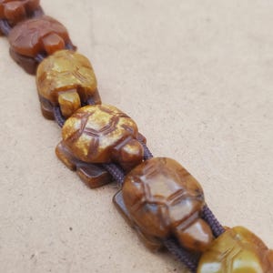 以下が含まれることがあります： 茶色のカメの形をしたビーズブレスレット。カメは明るい茶色の素材から彫られており、濃い茶色のコードに通されています。カメには詳細な甲羅の模様があり、一列に並んでいます。
