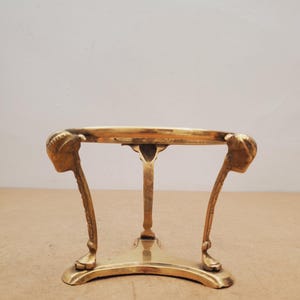 Puede incluir: Un soporte de latón vintage con una parte superior circular y tres patas. Las patas están adornadas con detalles de cabeza de carnero. El soporte está diseñado para sostener un objeto pequeño, como un cuenco decorativo o una maceta pequeña.
