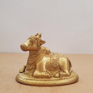 May include: A golden-colored brass sculpture of a reclining cow. The cow is adorned with detailed carvings, including a decorative saddle and headpiece. The sculpture is resting on a round, textured base, showcasing intricate patterns.