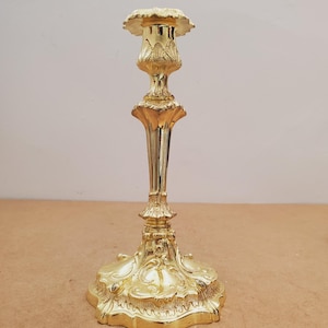Peut inclure: Un bougeoir doré avec un design floral détaillé. La base est large et ornée, soutenant une tige élancée qui mène à un dessus décoratif en forme de coupe. Le bougeoir est en métal et a une finition polie.