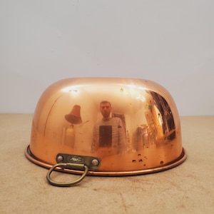 May include: A copper bowl with a rounded shape and a brass handle. The bowl reflects the surrounding environment, showing a person and other objects. The bowl is sitting on a light brown surface.