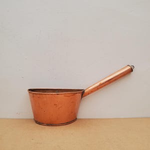 May include: A polished copper ladle with a wide, deep bowl and a long, straight cylindrical handle. The metal surface displays a warm, metallic orange hue with subtle variations in texture and tone, reflecting light across its curved exterior.