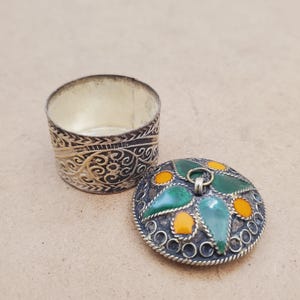 May include: An open, silver-colored container with an ornate, engraved design on the side. The lid is decorated with green and orange stones set in a silver frame. The lid has a small loop for opening.