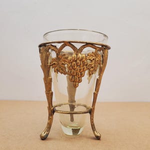 May include: A clear glass vase with a gold-coloured metal grape vine design around the top. The vase is supported by a gold-coloured metal stand with three curved legs. The vase is empty.