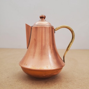 May include: A copper teapot with a brass handle and a matching lid. The teapot has a rounded base and a slender spout. The handle curves from the top of the pot. The teapot is on a light brown surface.