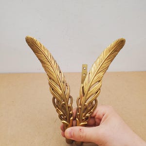 May include: Two ornate, gold-colored metal feather-shaped decorative elements. The feathers have detailed veining and are connected by a small, rectangular bracket with screw holes. The elements are held in a hand.