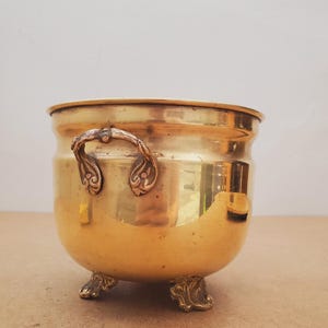 May include: A vintage brass planter with ornate handles and claw feet. The round planter has a polished, golden finish and a slightly curved body. The handles feature decorative scrollwork.