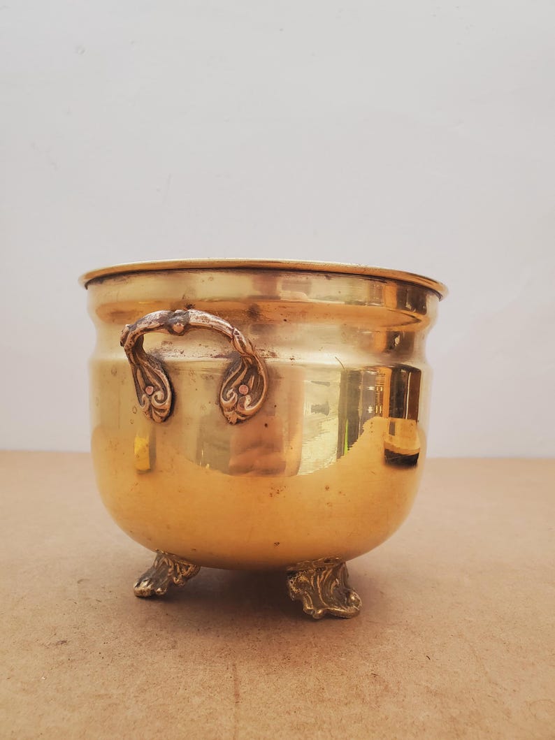 May include: A vintage brass planter with ornate handles and claw feet. The round planter has a polished, golden finish and a slightly curved body. The handles feature decorative scrollwork.