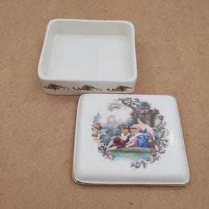 May include: A white porcelain trinket box with a square base and lid. The lid features a colourful illustration of two figures in a garden scene, surrounded by flowers. The box has a gold trim and floral accents.