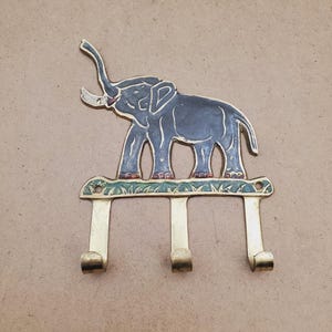 May include: A brass elephant-shaped wall hook with three hooks. The elephant is dark gray with white tusks and red accents. The base of the hook has a green and blue floral design.