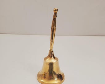Pode incluir: Um pequeno sino de latão dourado com uma alça. O sino tem uma forma arredondada e uma superfície lisa.