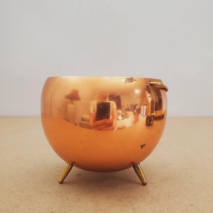 May include: A copper-colored, spherical bowl with three gold-colored legs and a small handle. The bowl's surface reflects the surrounding environment. The bowl is likely a decorative item or a small planter.