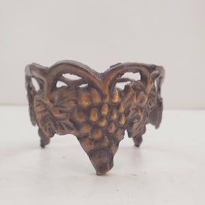 Puede incluir: Cuenco decorativo de color bronce con tres patas y diseño de racimo de uvas. El cuenco tiene un borde calado y un detallado motivo de uvas y hojas en la parte delantera. El cuenco es de metal y tiene un aspecto vintage.