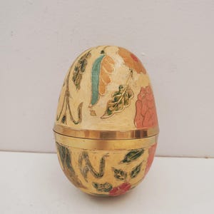 Könnte beinhalten: Eiförmige dekorative Box mit Scharnierdeckel. Die Box ist mit Blumen- und Blattmotiven in Orange-, Grün- und Rottönen auf cremefarbenem Hintergrund bemalt. Ein goldfarbenes Band umschließt die Mitte.