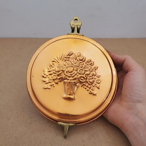 May include: A round, copper-colored wall hanging with a floral basket design. The metal piece has a hinged top and a small stand. The floral design is embossed on the front. The piece is held by a hand.