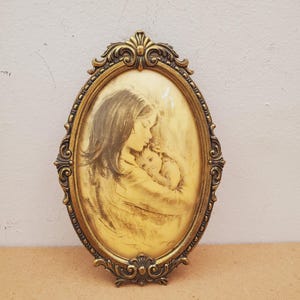 May include: An oval-shaped picture frame with an ornate, antique gold-colored finish. The frame contains a sepia-toned sketch depicting a person holding a small child close to their chest. The frame features intricate scrollwork and decorative detailing.