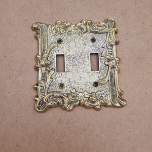 Placa de interruptor duplo vintage dos anos 1960, prateada/dourada e branca com estampa floral desgastada.
