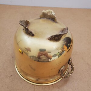 May include: A shiny brass bowl with three decorative feet and a handle. The bowl has a rounded shape and a polished surface, reflecting the surroundings. The handle is ornate, adding to the vintage aesthetic.