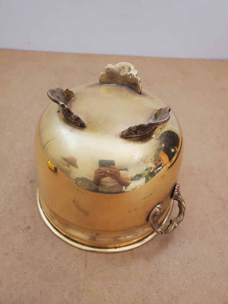 May include: A shiny brass bowl with three decorative feet and a handle. The bowl has a rounded shape and a polished surface, reflecting the surroundings. The handle is ornate, adding to the vintage aesthetic.