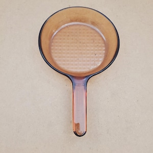 May include: A round, amber-coloured glass frying pan with a long handle. The interior base of the pan features a distinct, raised grid or waffle-like texture pattern. The glass is translucent, allowing light to pass through the warm brown material.