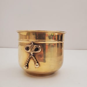May include: A small, gold-coloured brass planter with a decorative design on the side. The planter has a rounded shape and a smooth, shiny finish.