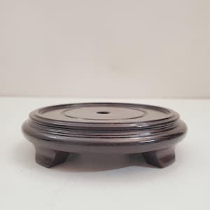Può includere: Un espositore in legno marrone scuro con una parte superiore circolare e tre gambe corte e curve. Il supporto ha un design a più livelli rialzato attorno alla parte superiore e un piccolo foro al centro. Il supporto è progettato per contenere un oggetto decorativo.