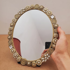 May include: Oval-shaped decorative mirror with a gold-colored frame. The frame is adorned with an intricate pattern of gold-colored floral and pearl-like embellishments. The mirror reflects the surrounding environment.