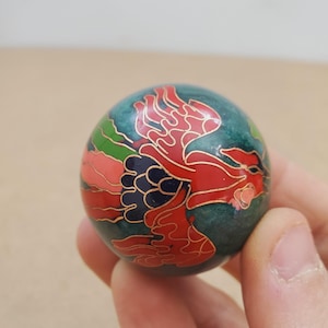 May include: A small, round decorative object with a teal base colour. The object features a red and gold floral design. The design is outlined in gold. The object is held in a person's hand.