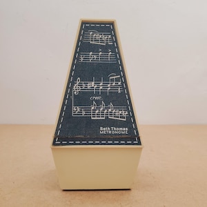 May include: A vintage Seth Thomas metronome. The beige base supports a dark blue, trapezoid-shaped top with musical notes and the word "cresc." printed on it. The metronome's name is printed on the front.