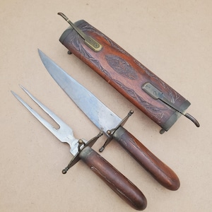 May include: A vintage carving set featuring a knife, fork, and sheath. The knife and fork have wooden handles and metal accents. The sheath is made of wood with decorative carvings and metal hardware. The set is likely from the early 20th century.