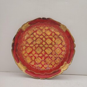K&ouml;nnte beinhalten: Ein rundes, rotes Holztablett mit einem goldenen geometrischen Muster. Das Tablett hat einen gewellten Rand.