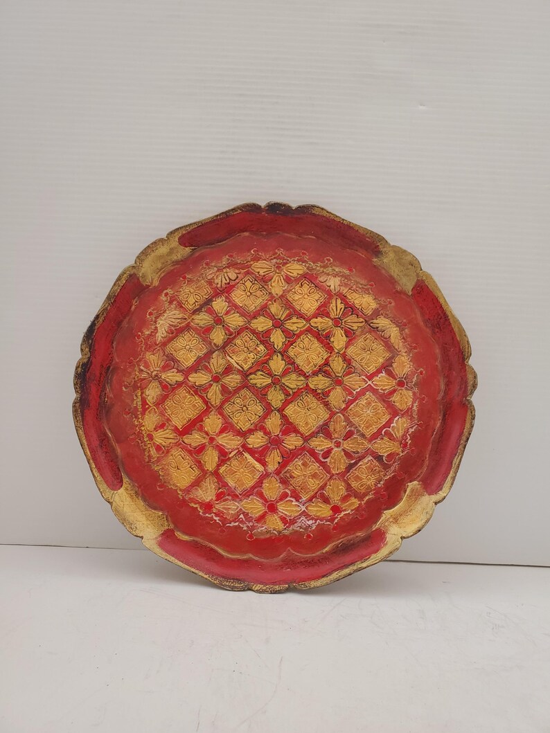 K&ouml;nnte beinhalten: Ein rundes, rotes Holztablett mit einem goldenen geometrischen Muster. Das Tablett hat einen gewellten Rand.