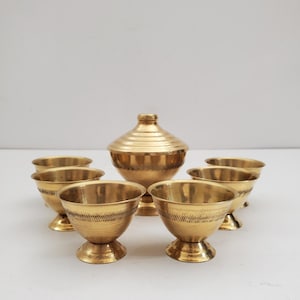 May include: A set of six small, gold-colored bowls with a matching lidded container. The bowls have a pedestal base and etched horizontal lines. The container has a rounded body and a conical lid.